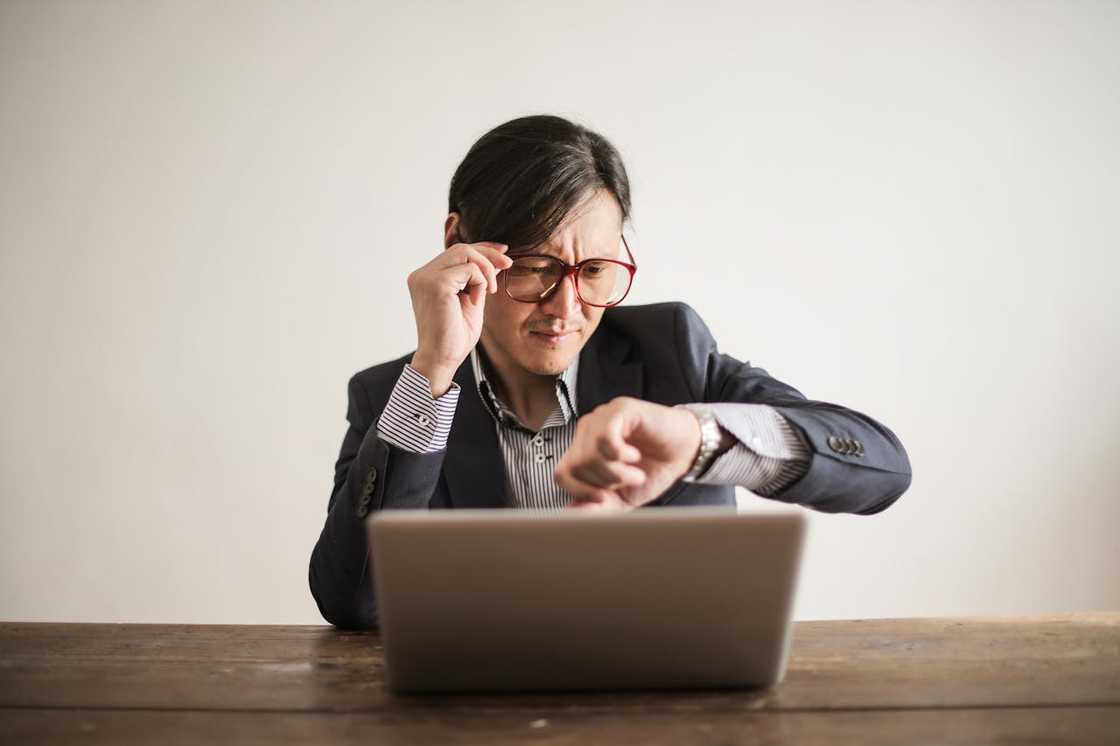 A man in a suit sits at a desk, looking at his watch in frustration. A man in a suit sits at a desk, looking at his watch in frustration.