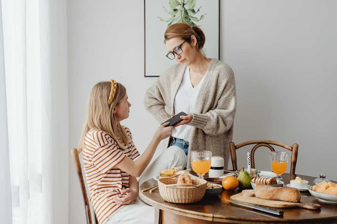 A woman stands by a dining table taking a phone from a seated girl. A woman stands by a dining table taking a phone from a seated girl.