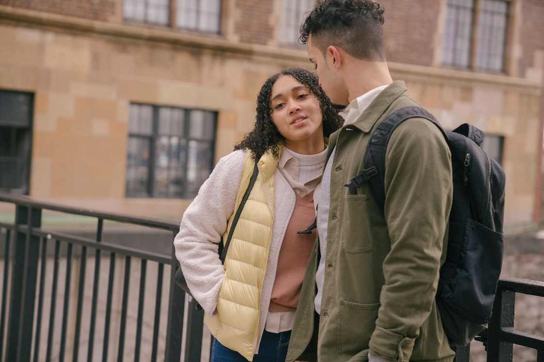 Two young adults stand close on a city walkway, quietly talking.