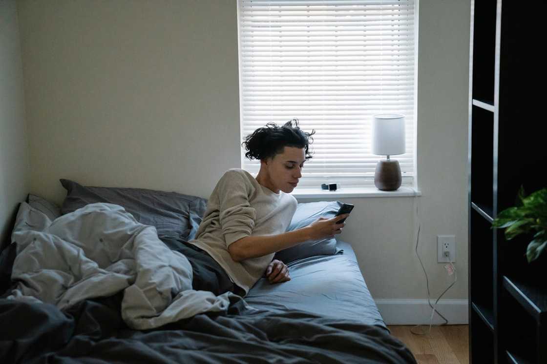 A person lies on a bed checking a phone near a window with closed blinds. A person lies on a bed checking a phone near a window with closed blinds.