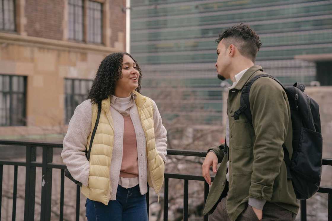Two young adults stand outdoors talking casually with city buildings in the background. Two young adults stand outdoors talking casually with city buildings in the background.