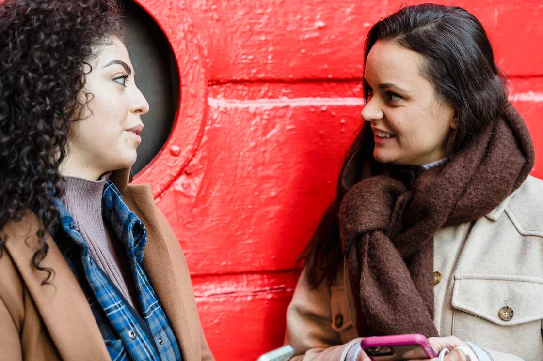 Two women standing close, smiling and talking beside a bright red wall. Two women standing close, smiling and talking beside a bright red wall.