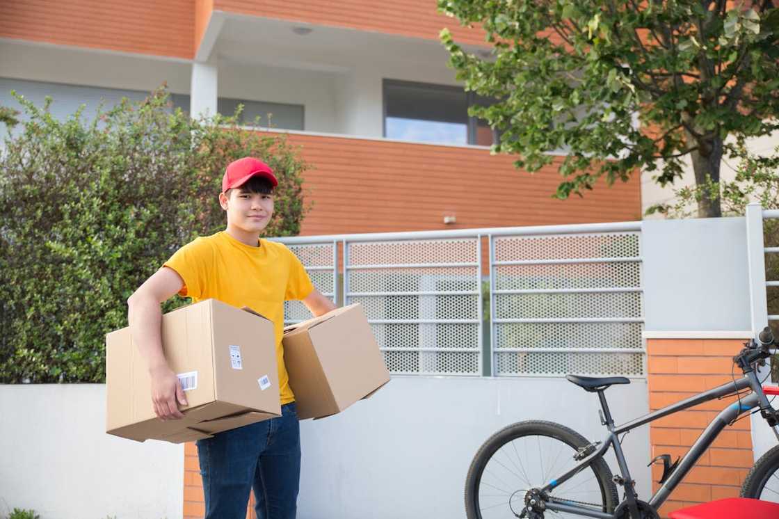 A young delivery worker stands outside a house holding two cardboard boxes, with a bicycle parked nearby. A young delivery worker stands outside a house holding two cardboard boxes, with a bicycle parked nearby.