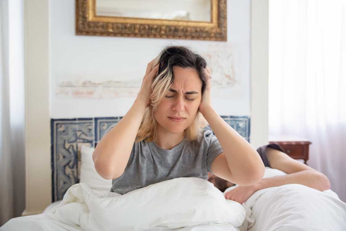 A woman in grey shirt holds her head while sitting on the bed.