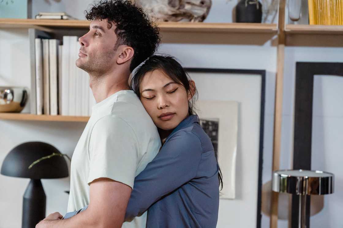 A woman hugs a man from behind indoors, resting her head against his back with her eyes closed. A woman hugs a man from behind indoors, resting her head against his back with her eyes closed.