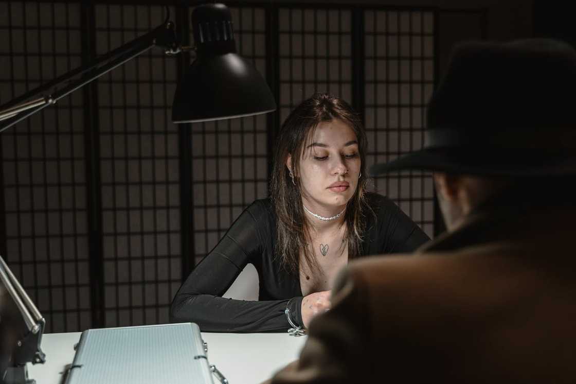 A woman sitting at a desk under a lamp, facing another person during questioning. A woman sitting at a desk under a lamp, facing another person during questioning.