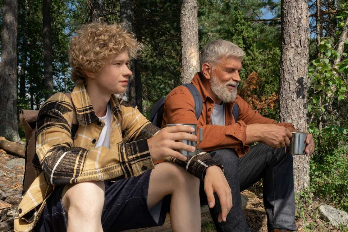 An older man and a young man are resting in a forest holding metal cups. An older man and a young man are resting in a forest holding metal cups.