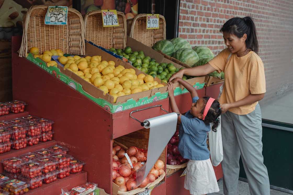 A woman helps a child reach for fruit while shopping at a market stall.