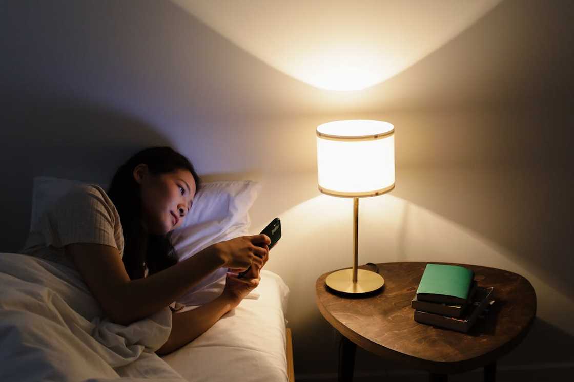 A woman lies in bed using her phone beside a lit bedside lamp at night. A woman lies in bed using her phone beside a lit bedside lamp at night.