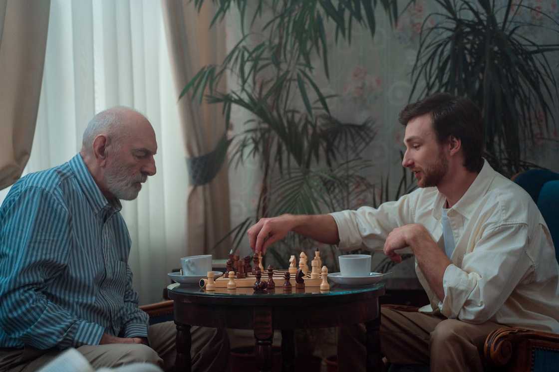 An older man and a younger man sit across a table playing chess. An older man and a younger man sit across a table playing chess.