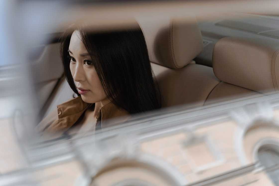 A person with long dark hair sits in the back seat of a car with beige leather interior.