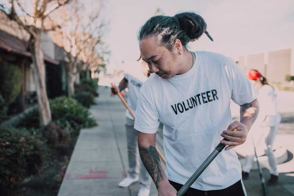A volunteer wearing a white shirt cleans a sidewalk outdoors while others work in the background.