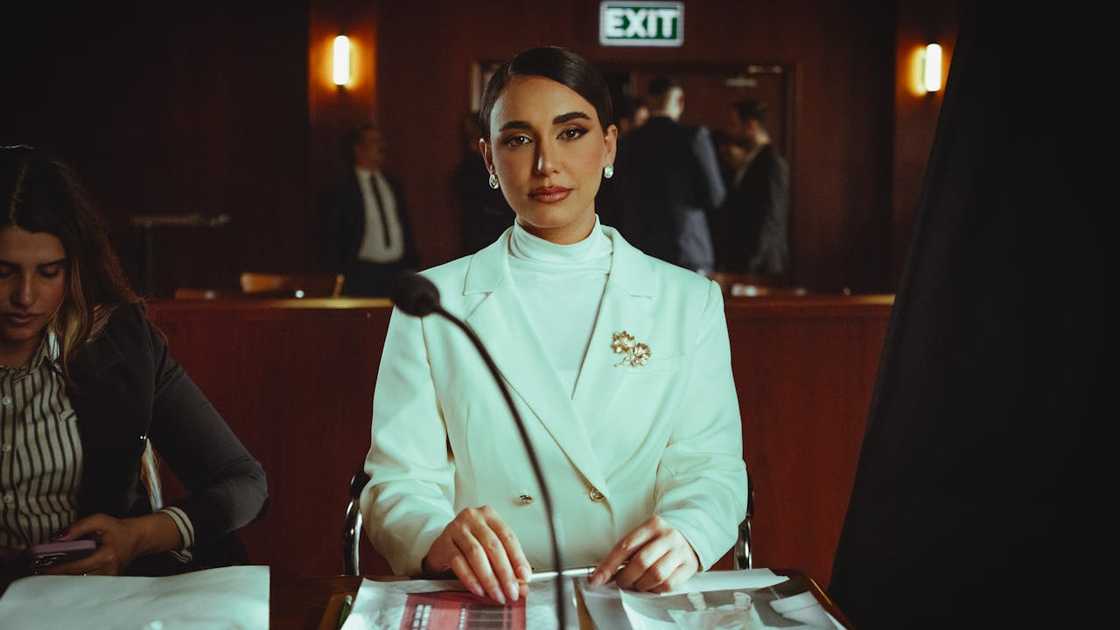 A woman in a white suit is seated at a courtroom desk, facing forward. A woman in a white suit is seated at a courtroom desk, facing forward.