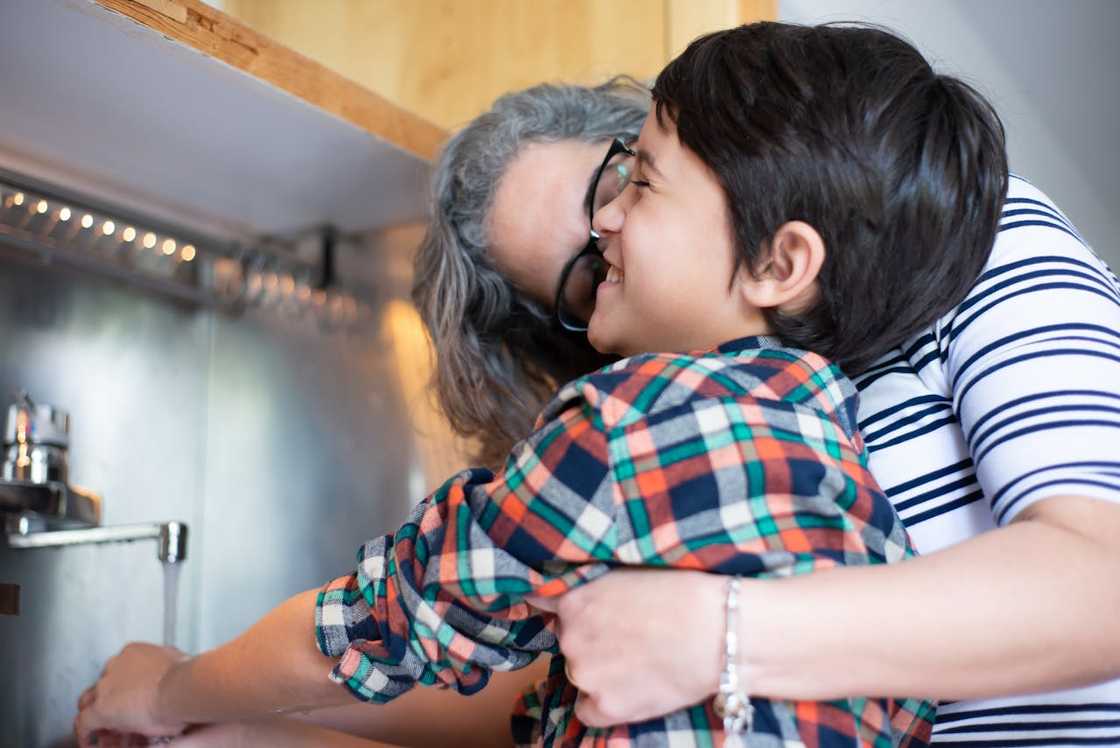 An adult hugs a smiling child while helping them wash their hands. An adult hugs a smiling child while helping them wash their hands.