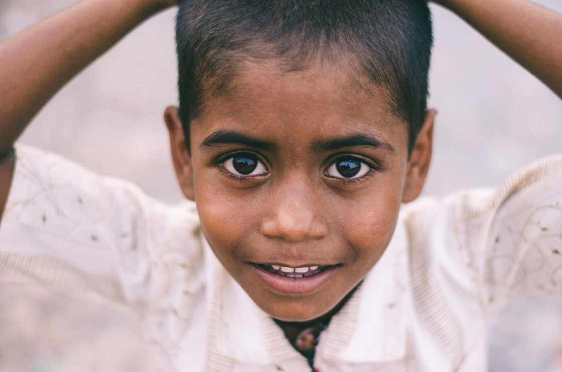 A close-up shot of a young boy in a white shirt holding his head outdoors.