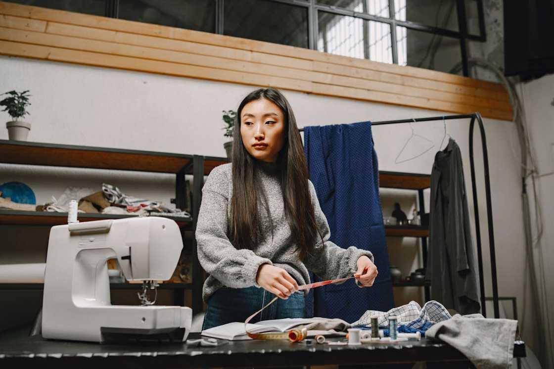 A woman measuring fabric beside a sewing machine in a small tailoring workspace. A woman measuring fabric beside a sewing machine in a small tailoring workspace.
