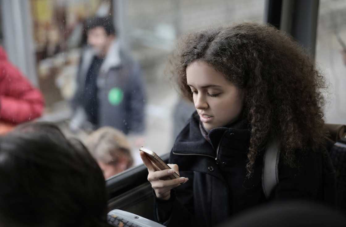 A young woman sits on public transport, looking at her phone while other passengers stand nearby.