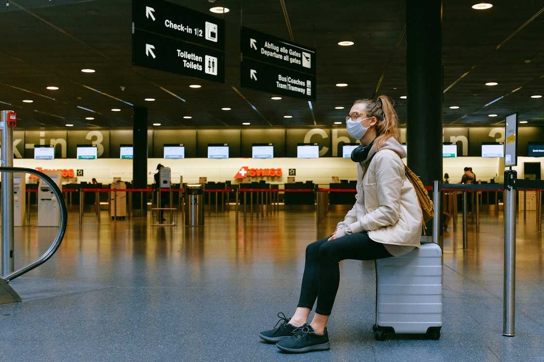 A person wearing a face mask sits on a suitcase in an airport terminal near check-in counters.