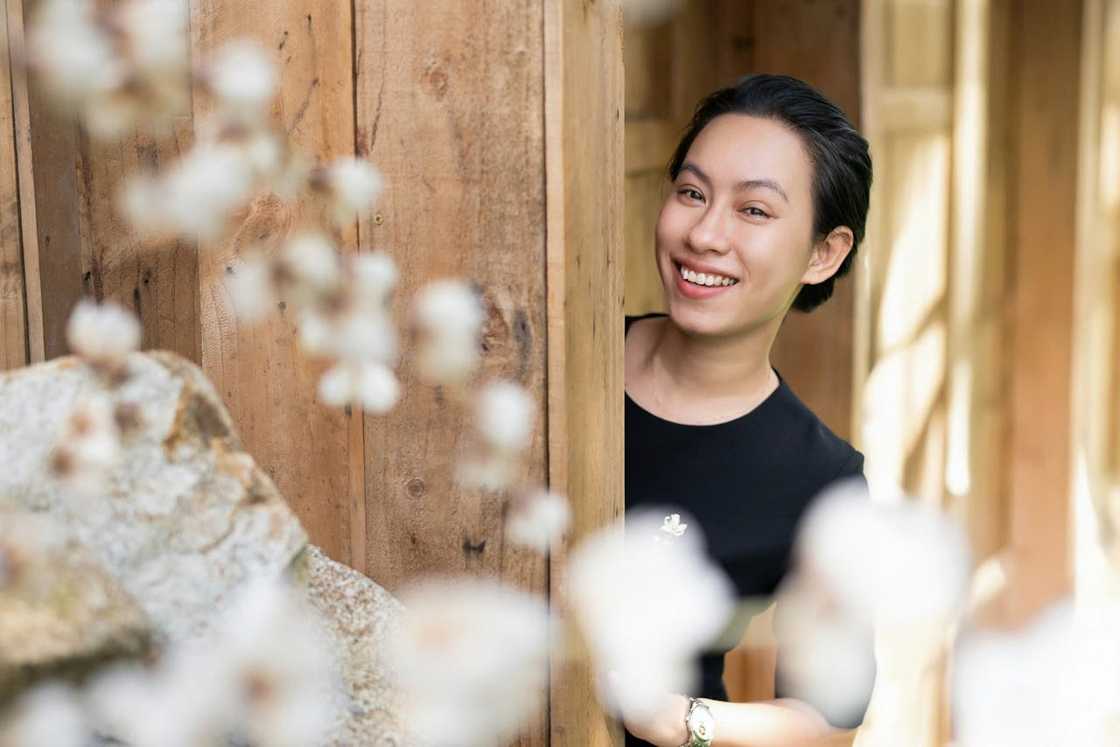 A woman smiles while standing partly behind a wooden wall, looking toward the camera. A woman smiles while standing partly behind a wooden wall, looking toward the camera.