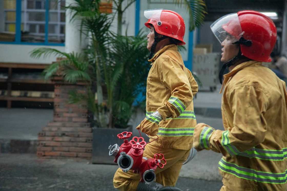 Two firefighters in yellow suits and red helmets work near a building. Two firefighters in yellow suits and red helmets work near a building.