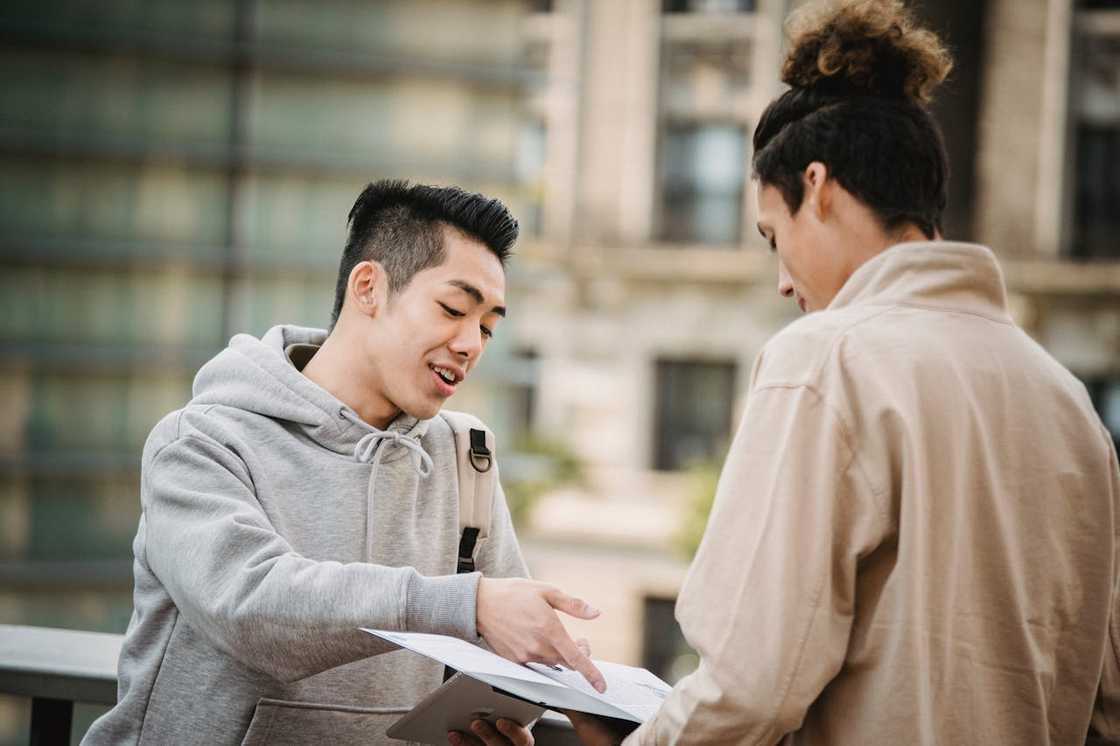 Two young adults standing outdoors, one pointing at a document while discussing it with the other.
