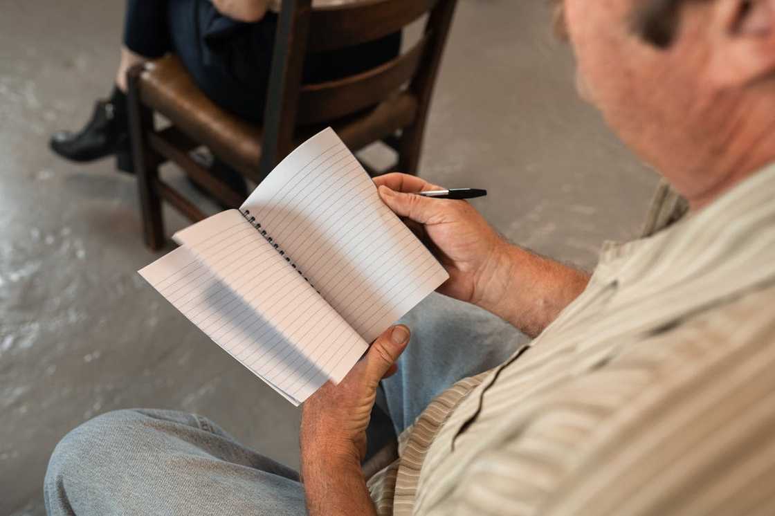 A man holds an open lined notebook and a pen while seated.