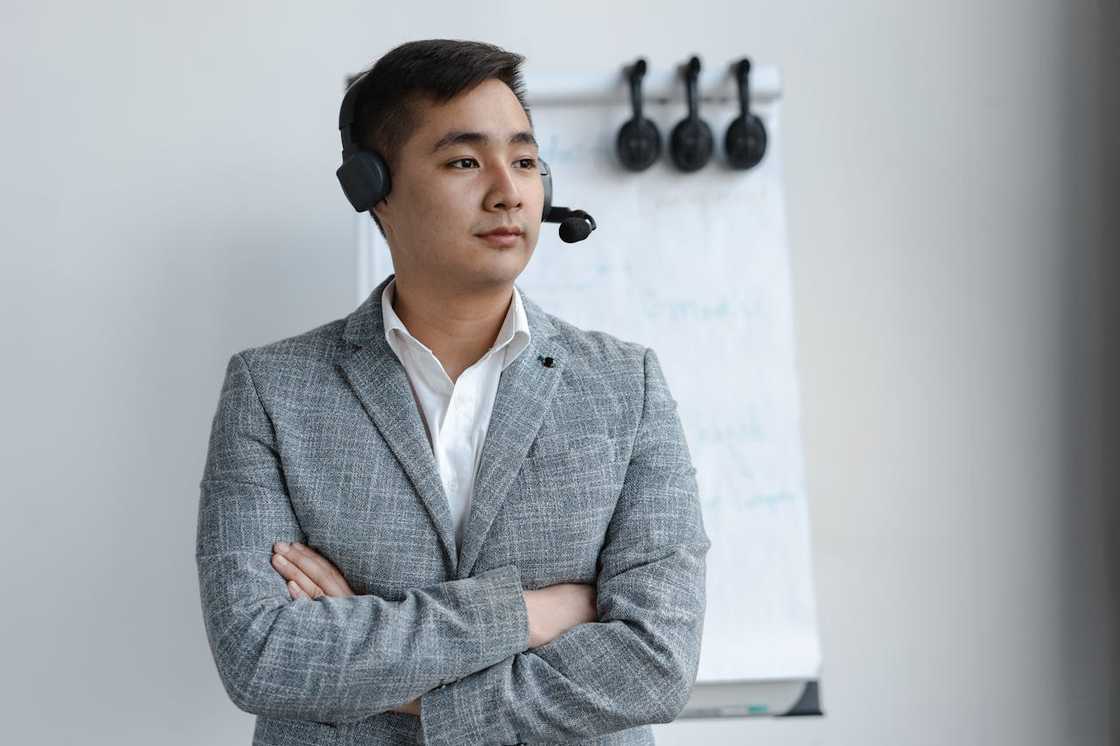 A man in a grey suit wearing a headset stands with his arms crossed in front of a flip chart.