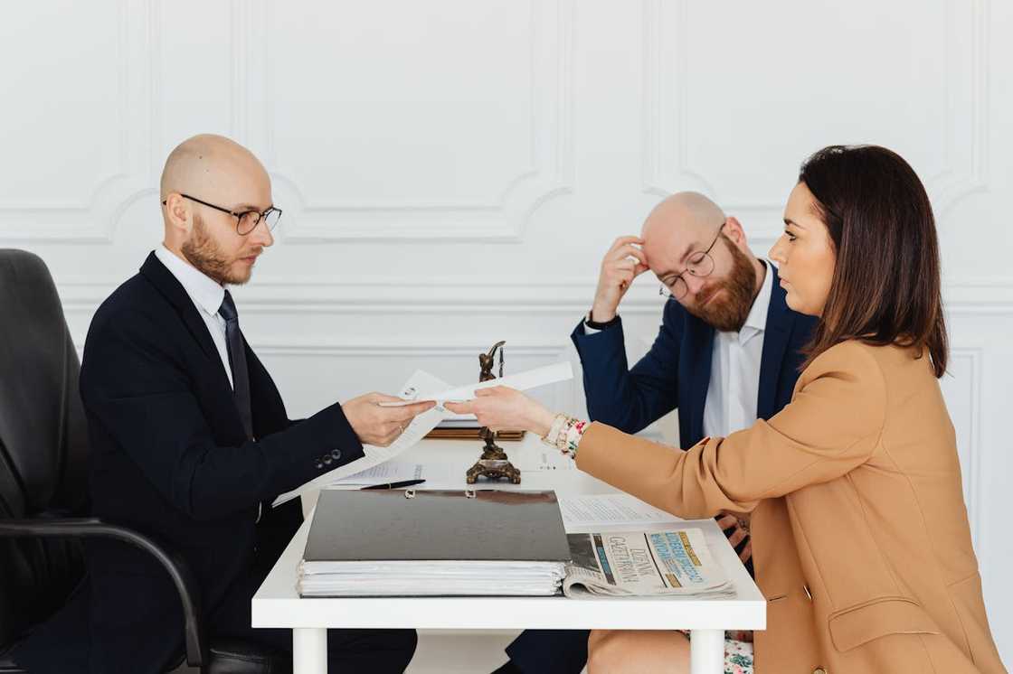 A woman hands documents to a lawyer across a desk while another man looks on anxiously. A woman hands documents to a lawyer across a desk while another man looks on anxiously.