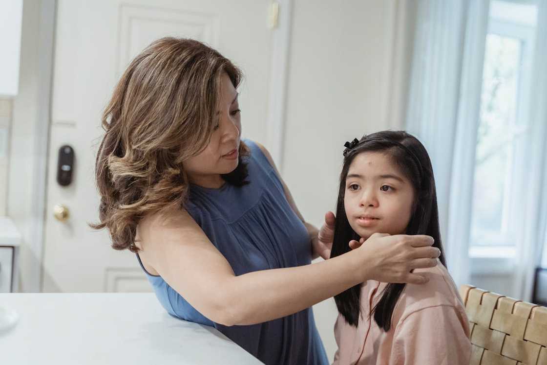 A woman gently adjusts a young girl’s hair while they sit together indoors. A woman gently adjusts a young girl’s hair while they sit together indoors.