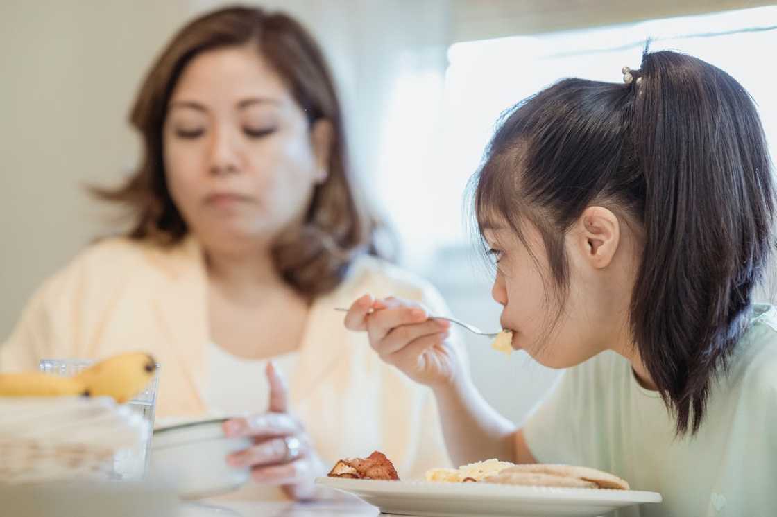 A young girl eats breakfast at a sunlit table with an adult.