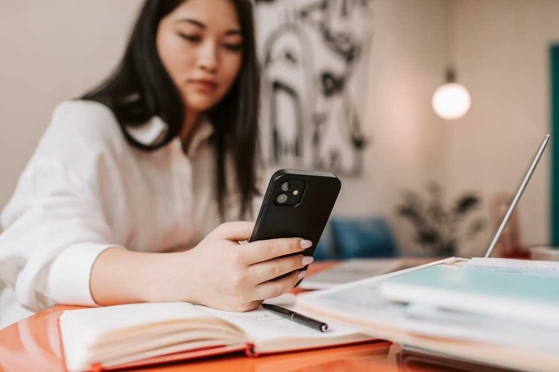 A woman sits at a desk looking at her smartphone next to books, papers, and a laptop. A woman sits at a desk looking at her smartphone next to books, papers, and a laptop.