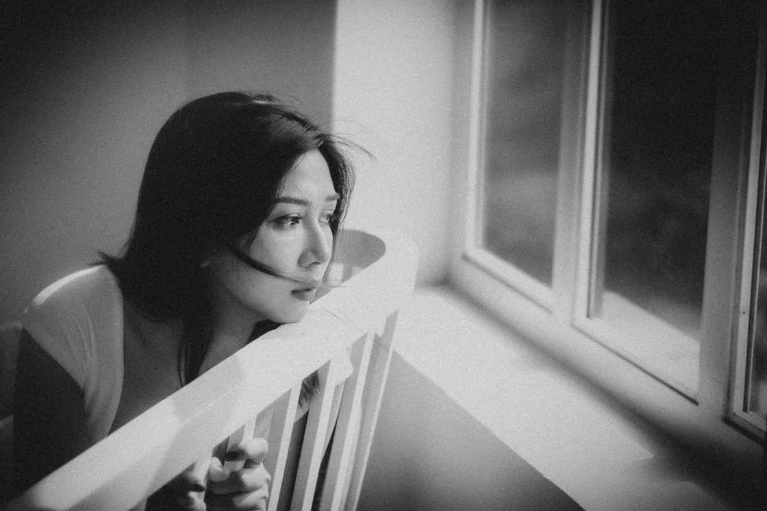 A woman leans on the back of a chair, gazing out of a window in quiet contemplation.