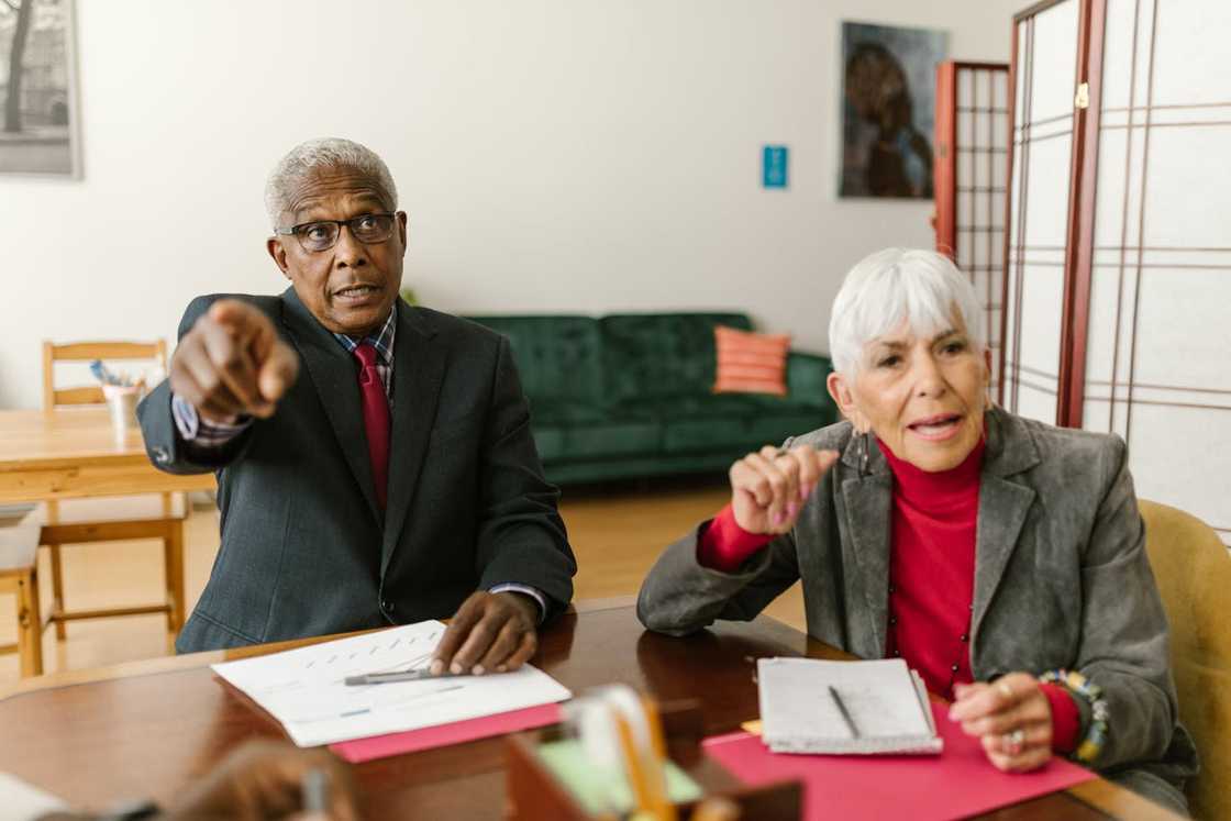 An older couple points and speaks during a serious meeting at a table.