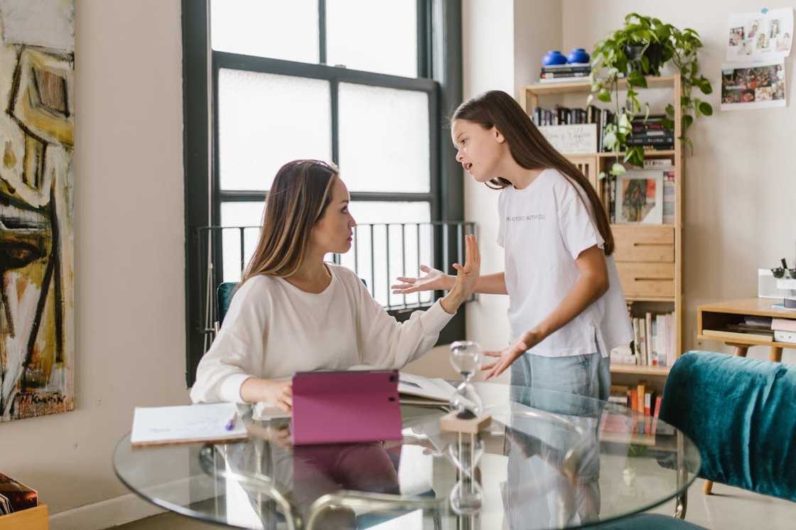 A woman sits at a glass table while a girl stands beside her, speaking with raised hands. A woman sits at a glass table while a girl stands beside her, speaking with raised hands.