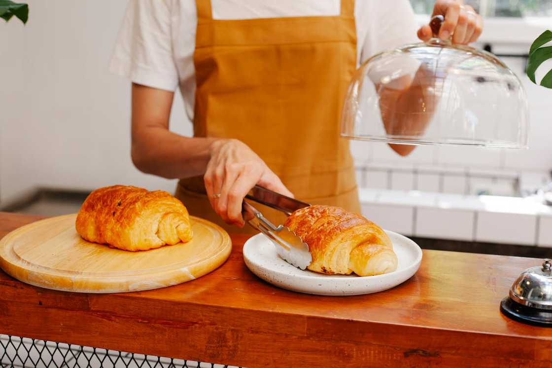A person in a mustard apron serves pastries at a café counter using tongs and a glass dome.