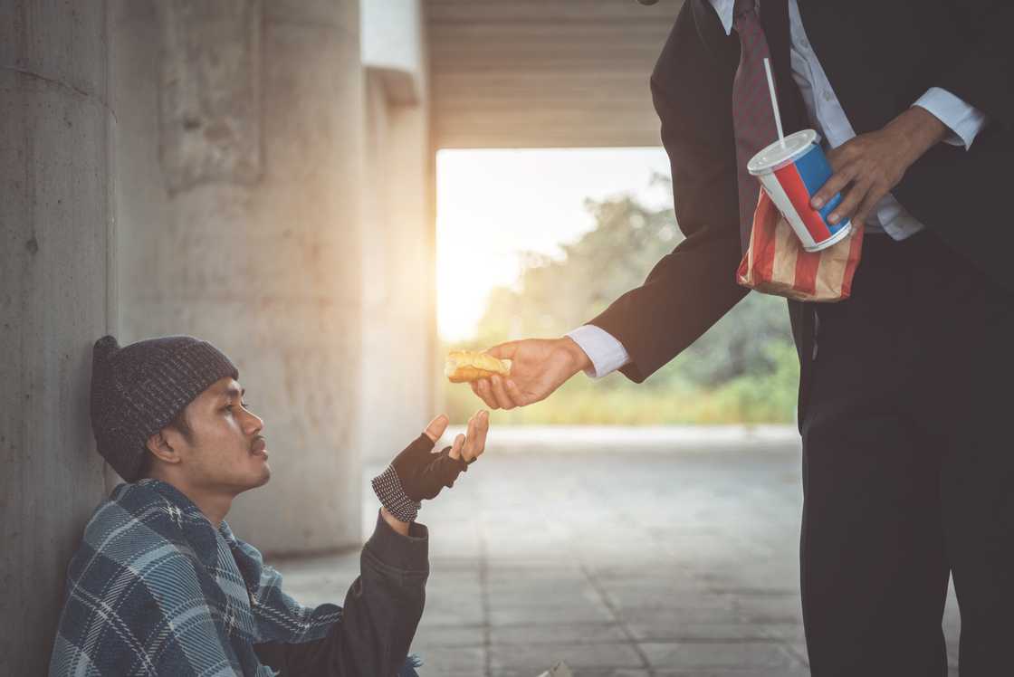 Person in a suit hands food to another individual sitting on the ground. Person in a suit hands food to another individual sitting on the ground.