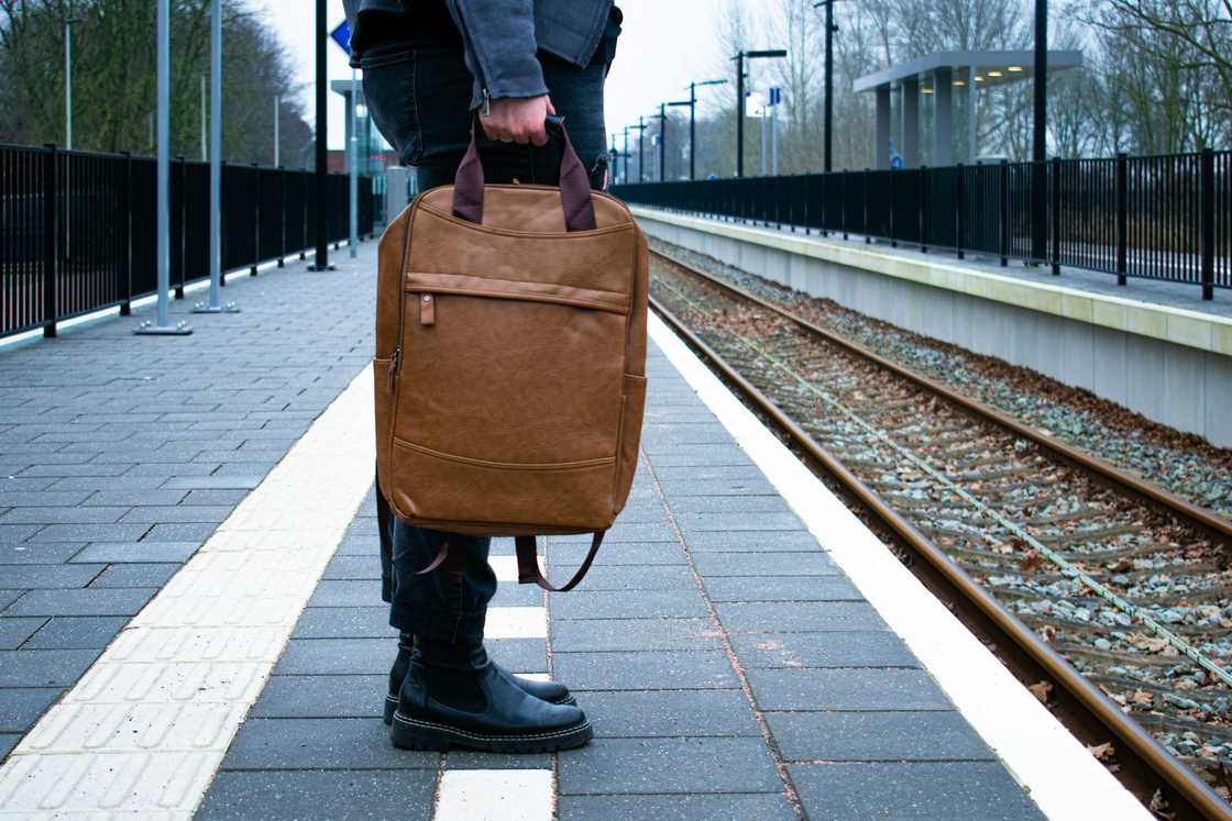 A person in dark clothes holds a brown backpack on a train platform with tactile paving.