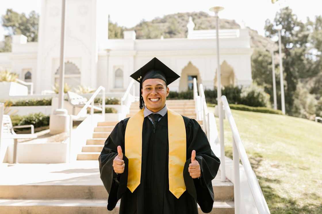 A graduate in a black gown, yellow stole, and mortarboard stands on outdoor steps. A graduate in a black gown, yellow stole, and mortarboard stands on outdoor steps.