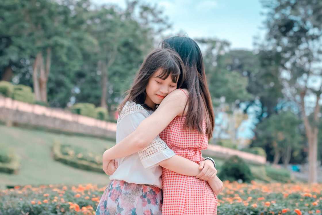 A mother and her teenage daughter hug in a garden.