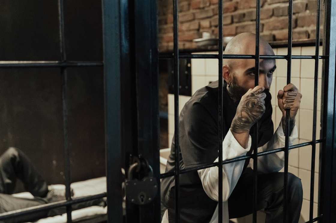 A man sitting behind prison bars, holding the metal rails inside a jail cell. A man sitting behind prison bars, holding the metal rails inside a jail cell.