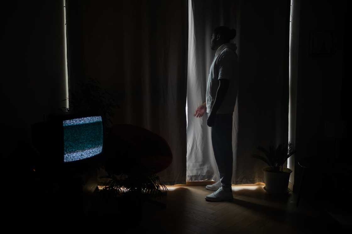 A man stands by a window looking out through partially opened curtains.