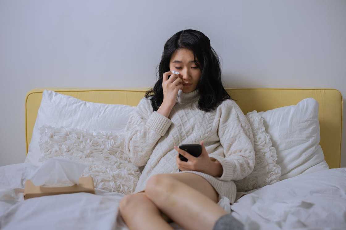 A woman in a cream sweater sits on a bed, holding a tissue and looking at a smartphone.