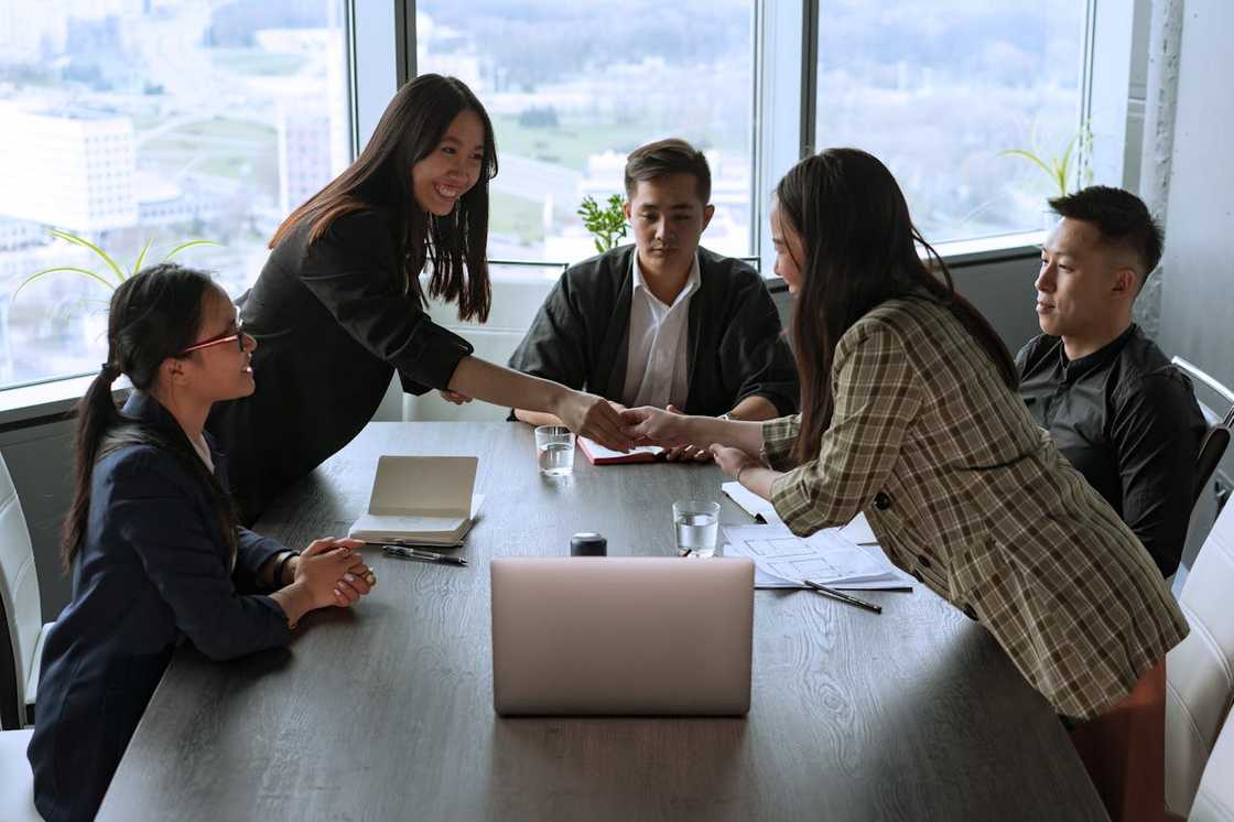 Five professionals sit around a conference table with two standing and shaking hands across it.