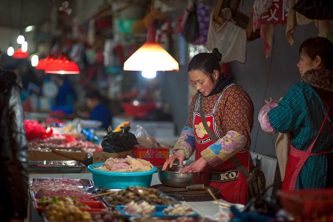 A woman in a red apron prepares food at a busy indoor market stall.