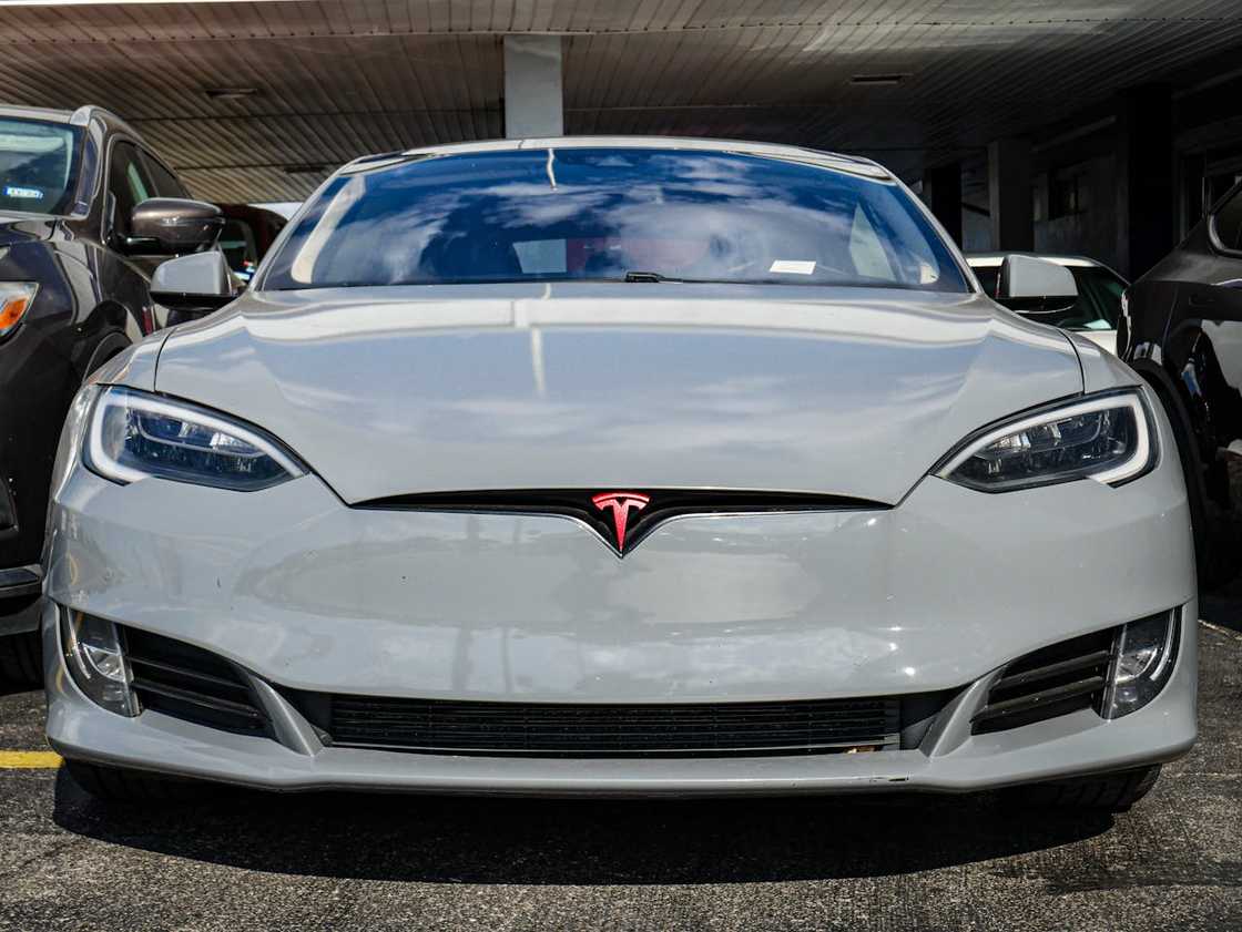 Front view of a silver Tesla parked in a lot between other vehicles, with red Tesla logo on the grille. Front view of a silver Tesla parked in a lot between other vehicles, with red Tesla logo on the grille.