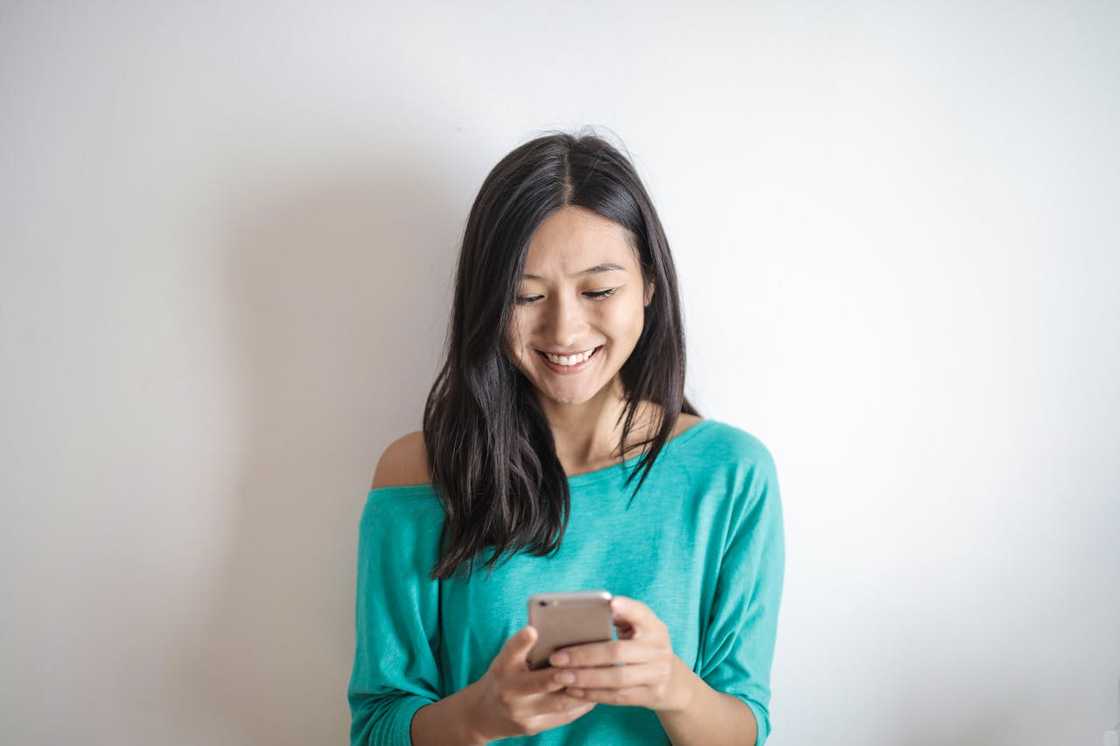 A woman stands against a plain wall, smiling as she looks down at her phone. A woman stands against a plain wall, smiling as she looks down at her phone.
