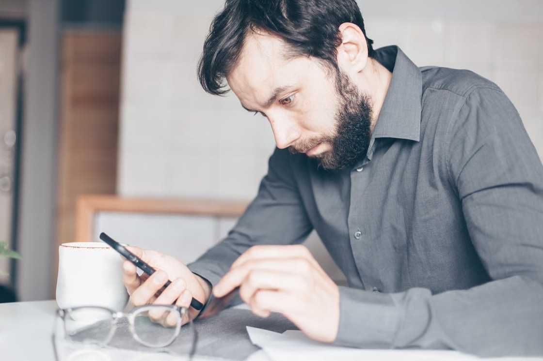 A man sitting at a table looks intently at his phone beside a cup and papers. A man sitting at a table looks intently at his phone beside a cup and papers.