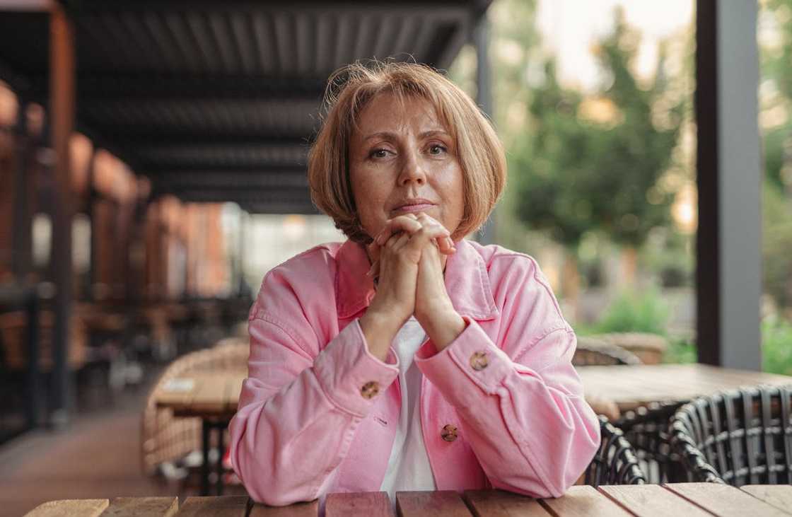 A woman sits at an outdoor table with her hands clasped, looking ahead thoughtfully. A woman sits at an outdoor table with her hands clasped, looking ahead thoughtfully.