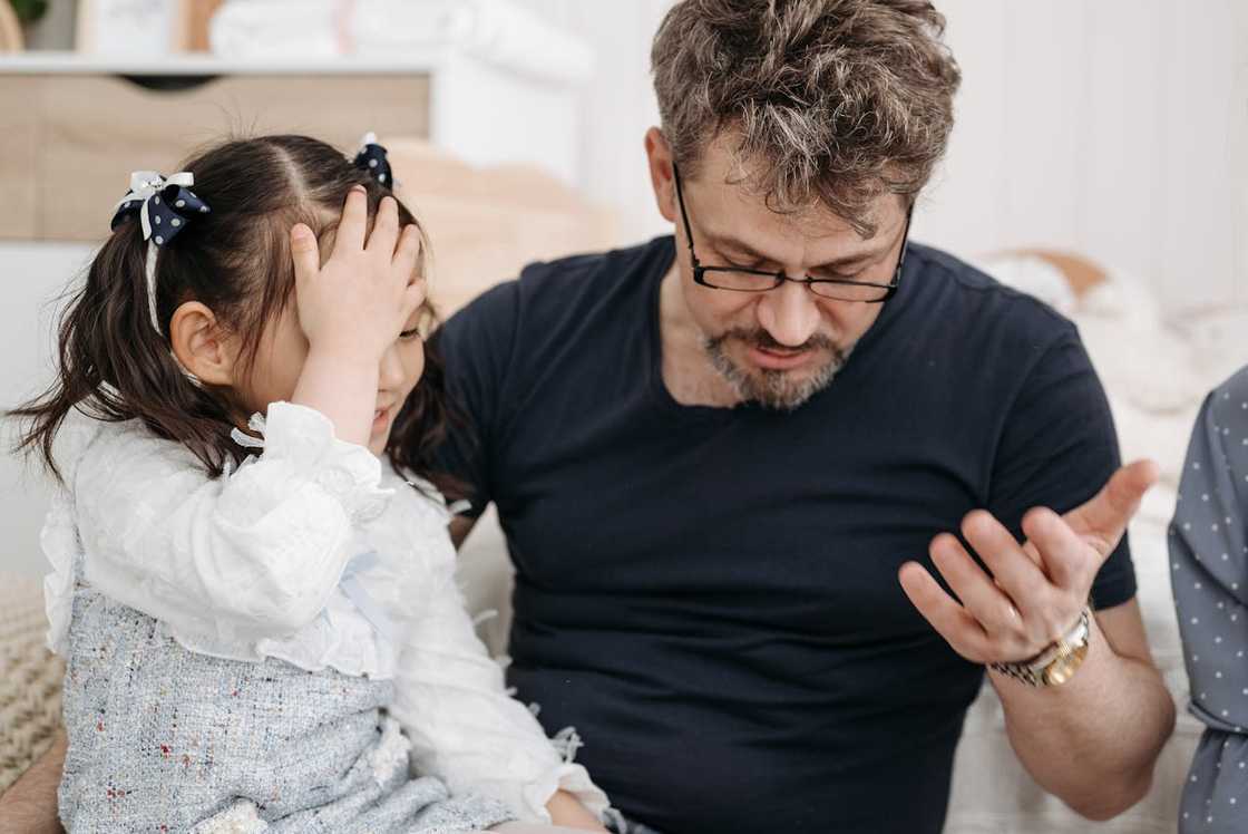 A man gestures while talking to a young girl sitting beside him indoors.