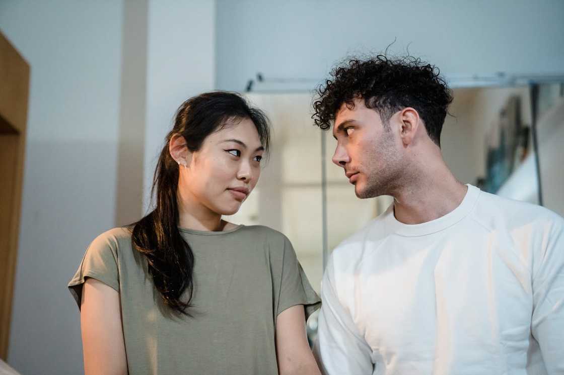 A couple standing indoors, looking at each other with serious expressions.